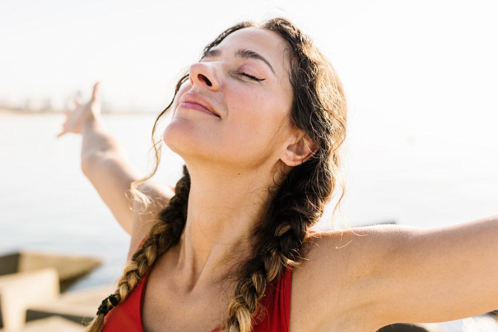 Young adult hispanic woman breathing fresh air standing on the beach at summer - Portrait of sportive hispanic female relaxing by the sea - Wellness and healthcare lifestyle concept